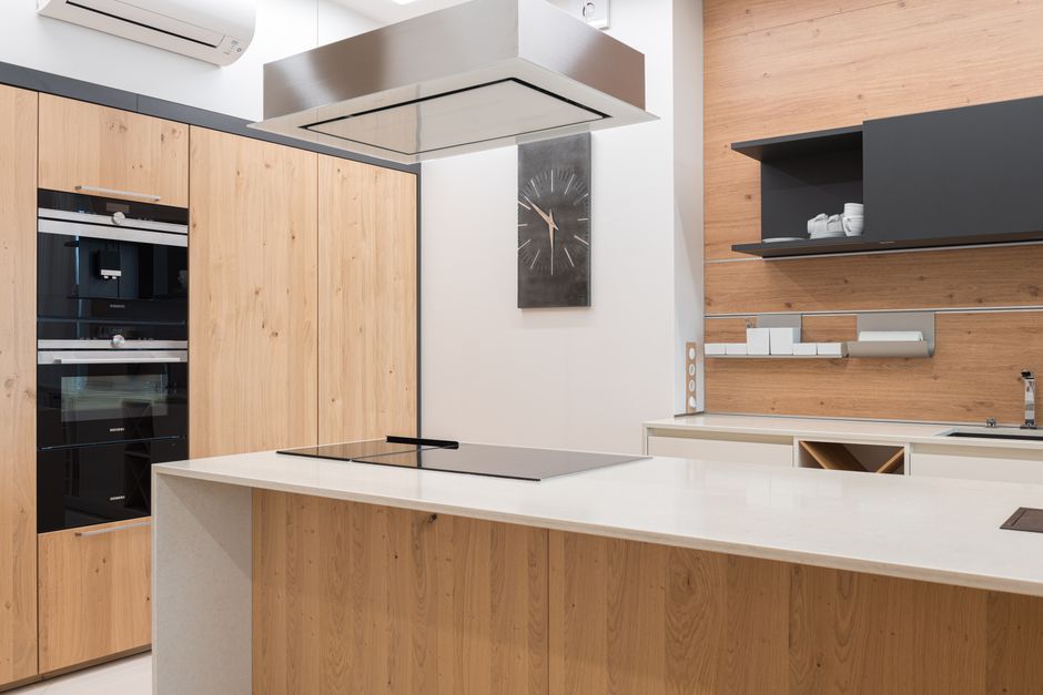A modern kitchen surface with a grey stone countertop, stainless steel oven, and white matte cabinets above and below. The oven door is closed, reflecting some light, with a control panel on top. The